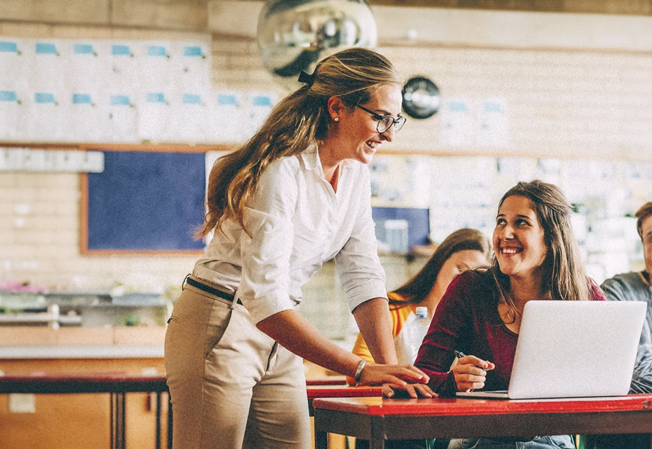 Female teacher helping female high school-aged student as she works on laptop at desk in classroom